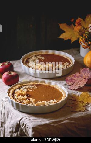 Traditional homemade autumn pumpkin pies for Thanksgiving or Halloween dinner served in ceramic dish with yellow autumn leaves, pumpkins and apples on linen table cloth. Dark rustic style Stock Photo