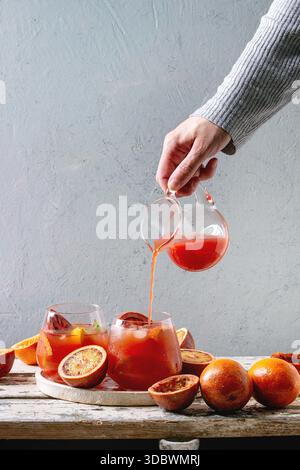 Female's hand poured from jug juice in blood orange iced cocktail in glasses, decorated by slice of oranges and fresh mint, served in ceramic tray on white wooden table with grey wall at background. Stock Photo