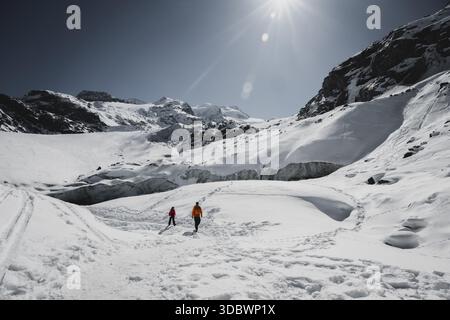 View of two people walking across the stark white snow under a bright sun in a mountainous landscape, Pontresina, Grisons Canton, Switzerland. Stock Photo
