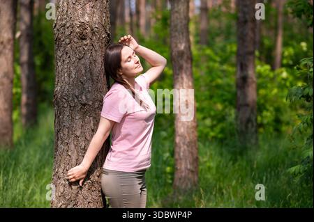 Young woman leaning against a tree in the forest, smiling and stretching her arms in the sun Stock Photo