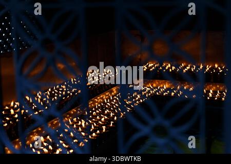 Rows of glowing temple oil lamps seen through a decorative metal grille, creating an abstract background of warm fire light and spiritual atmosphere. Stock Photo
