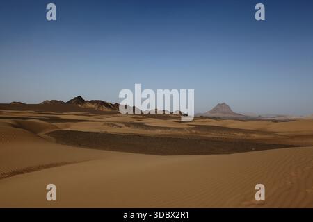 View of stark desert landscape with undulating sand dunes meeting distant dark, rugged mountains beneath a clear blue sky, Noshki, Balochistan, Pakist Stock Photo