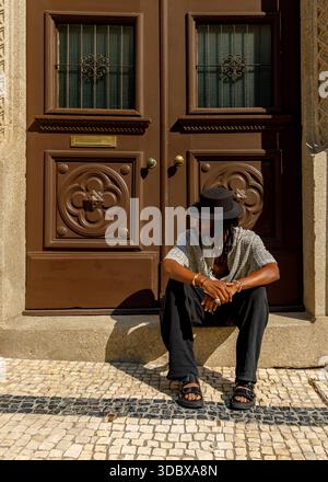 A man wearing a hat and casual clothing sits on the steps in front of a large brown door. Stock Photo