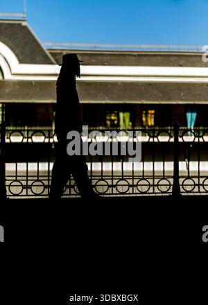 A silhouetted figure wearing a hat walks in front of a building with a wrought iron fence. Stock Photo