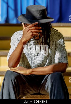A person wearing a black hat and a white shirt is sitting on the stairs with their hand covering their face. Stock Photo