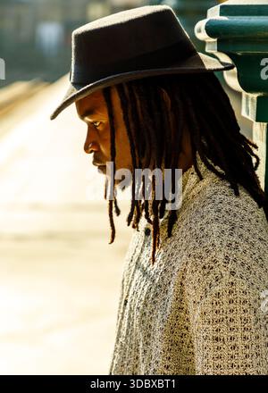 A man with dreadlocks wearing a hat and a sweater, leaning against a green structure. Stock Photo