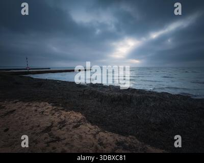 A wide-angle landscape of a moody, overcast sky with sunbeams breaking through dark clouds over the Oresund sea. The foreground features the sandy sho Stock Photo