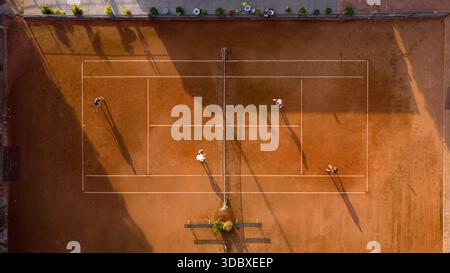 Aerial view of players on a terracotta tennis court, bathed in golden sunlight, casting long shadows across the meticulously lined field, Sint Maarten Stock Photo