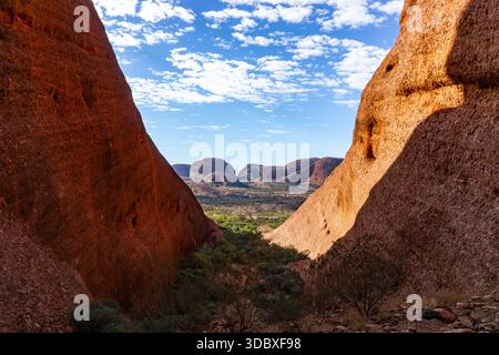 View of sunlit, colossal red rock formations framing a vibrant green valley under a bright blue, cloud-strewn sky, Kata Tjuta - Valley of the Winds, N Stock Photo