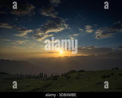 View of the sun setting behind hazy mountains casting long shadows over green hills and scattered trees, Sri Paye, Khyber Pakhtunkhwa, Pakistan. Stock Photo