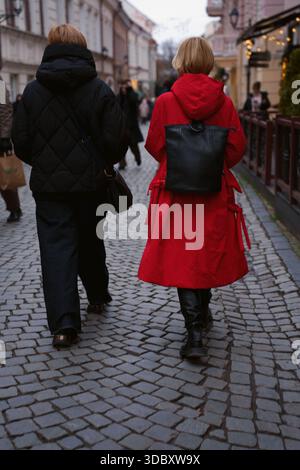two people stroll down a cobblestone street in a historic urban setting, wearing black and red coats, with late afternoon light casting soft shadows a Stock Photo