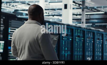 Engineer walking in server farm, automating system diagnostics using machine learning predictions. IT specialist working in data center using notebook to configure gear for AI tasks, camera B Stock Photo