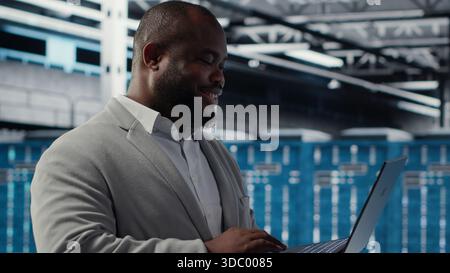 Data center engineer deploying AI driven management tools using laptop. Server farm employee inspecting server clusters, updating network topologies using artificial intelligence, camera B Stock Photo