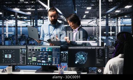 Data center manager supervising developer using device to monitor neural network AI LLMs. Team leader checking progress done by worker overseeing servers powering machine learning processes, camera A Stock Photo