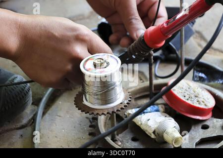 Close up of concentrated repairman hand soldering wire on electronic component with red soldering iron and silver spool of lead wire in messy workshop Stock Photo