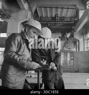 Sloviansk, Ukrainian SSR, USSR, 1985. An archival photograph taken at the AIZ plant (Armaturno-Izolyatorny Zavod). Two employees in construction helmets, likely including Agatuev, discuss production tasks in the workshop. The senior specialist in a leather jacket checks notes in a notebook held by his younger colleague. The background shows the scale of the industrial facility with cranes and equipment. A vivid episode of the working process in the peaceful Donbas during the Soviet era. Stock Photo