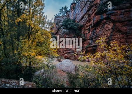 River flowing past steep red sandstone cliffs with yellow autumn foliage and boulders under a cloudy sky Stock Photo