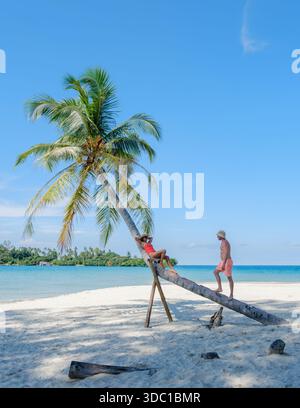 Two friends enjoy a sunny day at Koh Kood Island, Thailand, swinging from a palm tree and laughing together on soft white sand. The turquoise ocean ad Stock Photo