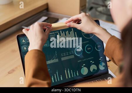 Young adult woman using laptop computer, analyzing digital dashboard with business analytics charts and graphs, hands interacting with sticky note on screen Stock Photo