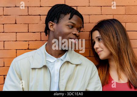 Young multi ethnic couple stands close together by a brick wall, gazing into each other's eyes with love and affection Stock Photo
