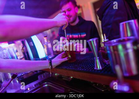 Rovigo, italy 18 december 2025: barman hands stirring a vibrant purple cocktail in a glass, serving a customer under colorful neon lights during night Stock Photo