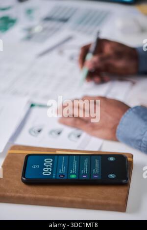 Man working at desk with smartphone displaying notifications in foreground, hands holding pen and reviewing printed documents with charts and diagrams Stock Photo