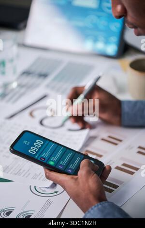 Young adult Black man holding smartphone with notifications visible while analyzing printed charts and graphs at desk, multitasking with pen in hand and digital device in workspace Stock Photo