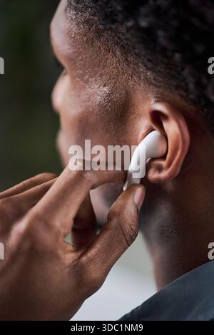 Black young adult man adjusting wireless earbud in ear, closeup of hand and side of face, focusing on technology use and personal audio device, blurred background Stock Photo