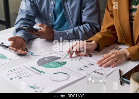 Caucasian woman and Black man analyzing financial charts and graphs at desk, discussing business strategy while reviewing printed reports and using smartphone during meeting Stock Photo