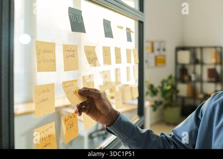 Black man organizing sticky notes on glass wall, managing project tasks in modern office setting, hand reaching toward notes with to do list visible, focusing on workflow planning Stock Photo