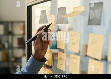 Black man writing on sticky notes attached to glass wall, organizing tasks and planning project workflow, hand holding marker, visible notes with various work-related reminders and goals Stock Photo
