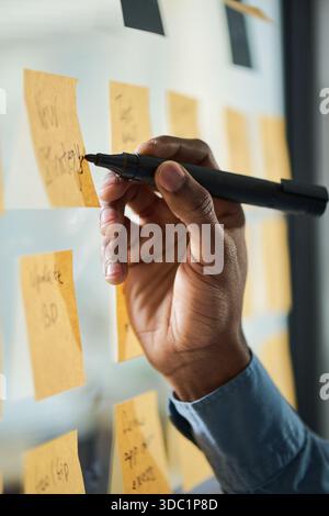 Black man hand writing on sticky notes attached to glass wall, planning or organizing tasks in office setting, visible wrist and part of sleeve suggesting young adult professional Stock Photo