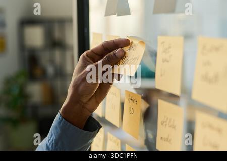 Black man organizing sticky notes on glass wall, hand placing note labeled new strategy among various reminders, business planning or brainstorming session in modern office setting Stock Photo