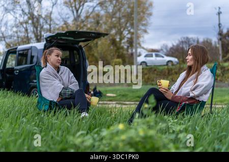Friends traveling conversation women relax drinking coffee sitting in camping chairs beside van Stock Photo