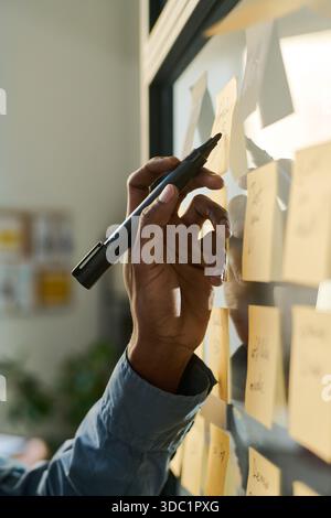Black young adult man writing on sticky notes attached to glass wall, hand holding marker in focus, brainstorming ideas during collaborative planning session in modern office setting Stock Photo