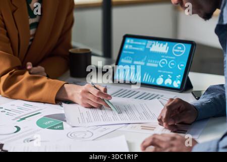 Caucasian young adult woman and Black man analyzing financial documents and discussing data with digital tablet displaying charts and graphs on office desk Stock Photo