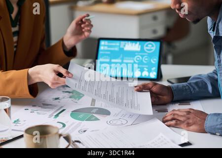 Caucasian woman and Black man analyzing printed business reports and discussing data at desk with digital tablet displaying charts, focusing on teamwork and financial planning Stock Photo