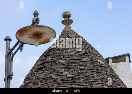 close-up of the ancient conical stone roof of a trullo, topped with a pinnaculum, juxtaposed with an old street lamp and a faint crescent moon in the Stock Photo