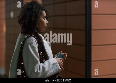 Young adult Black woman standing in profile holding smartphone, waiting near modern wall, preparing for date, curly hair visible, focused expression, evening setting Stock Photo
