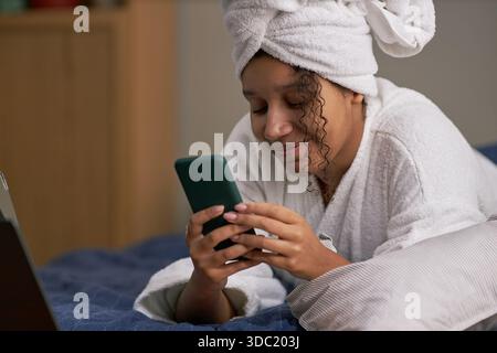 Young adult Black woman lying on bed smiling while using smartphone, wearing bathrobe and towel on head, engaging in online dating or chatting, laptop partially visible nearby Stock Photo
