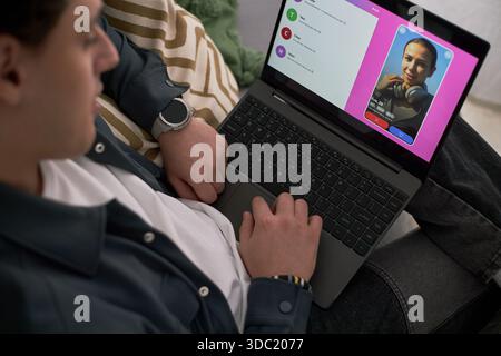 Young adult Caucasian man using laptop while browsing dating app, viewing profile of young adult Black woman smiling at camera, relaxing on couch, hand resting on keyboard Stock Photo