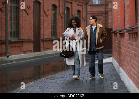 Young adult Black woman holding bouquet of flowers, walking beside young adult Caucasian man on city street, smiling and talking during romantic date in urban setting Stock Photo