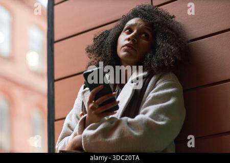 Young adult Black woman standing outdoors holding smartphone, looking up thoughtfully, leaning against wooden wall, using dating app, curly hair visible, urban background blurred Stock Photo