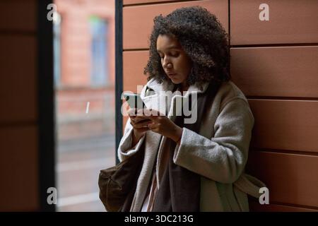 Young adult Black woman standing outdoors using smartphone, looking at screen with focused expression, holding device with both hands, engaging in online dating communication Stock Photo