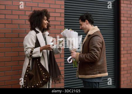 Young adult Caucasian man smiling while giving bouquet of flowers to young adult Black woman holding coffee cup outdoors, both standing near brick wall, dating context Stock Photo