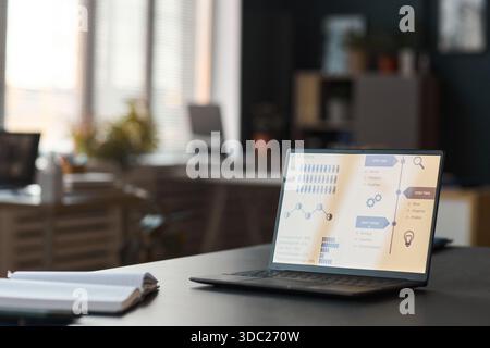 Laptop displaying business analytics dashboard with charts and workflow diagrams on desk in modern office, sunlight streaming through window, notebook and pen lying nearby Stock Photo