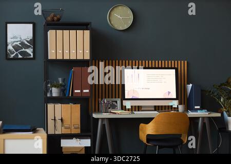Modern office workspace featuring computer monitor displaying business charts and graphs on desk with organized folders and binders on shelves against dark wall background Stock Photo