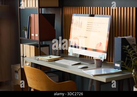 Modern workspace featuring desktop computer displaying business analytics charts and graphs on screen, organized desk with office supplies, shelving unit with folders and documents in background Stock Photo