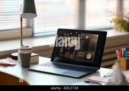 Open laptop displaying data analytics dashboard with world map and charts on screen, sitting on office desk near reusable coffee cup, lamp, and stationery, blinds covering window Stock Photo