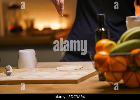 Rolled dough home kitchen cozy lifestyle moment. Unrecognizable person sprinkling flour above flattened pastry surface, warm evening light, calm domes Stock Photo